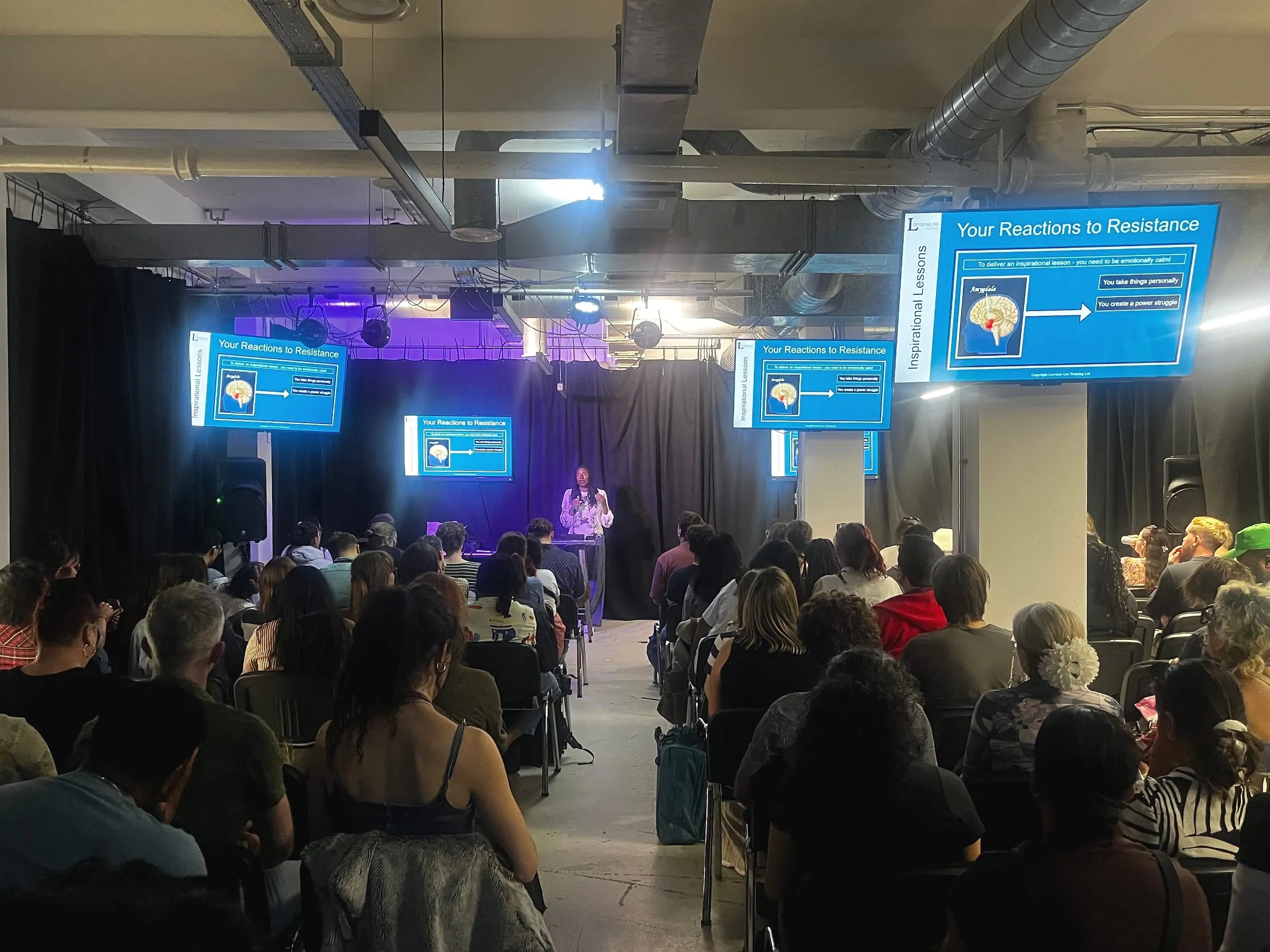 Lorraine Lee speaking from a stage to a full room, beside screens reading Your Reactions to Resistance
