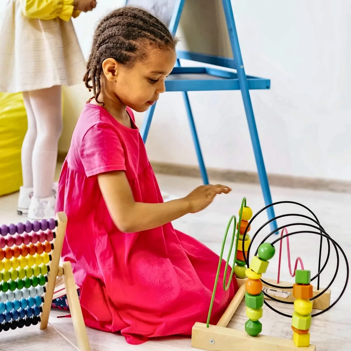A young girl in a pink dress sitting on the floor of a nursery, playing with a wooden bead maze beside a colourful abacus
