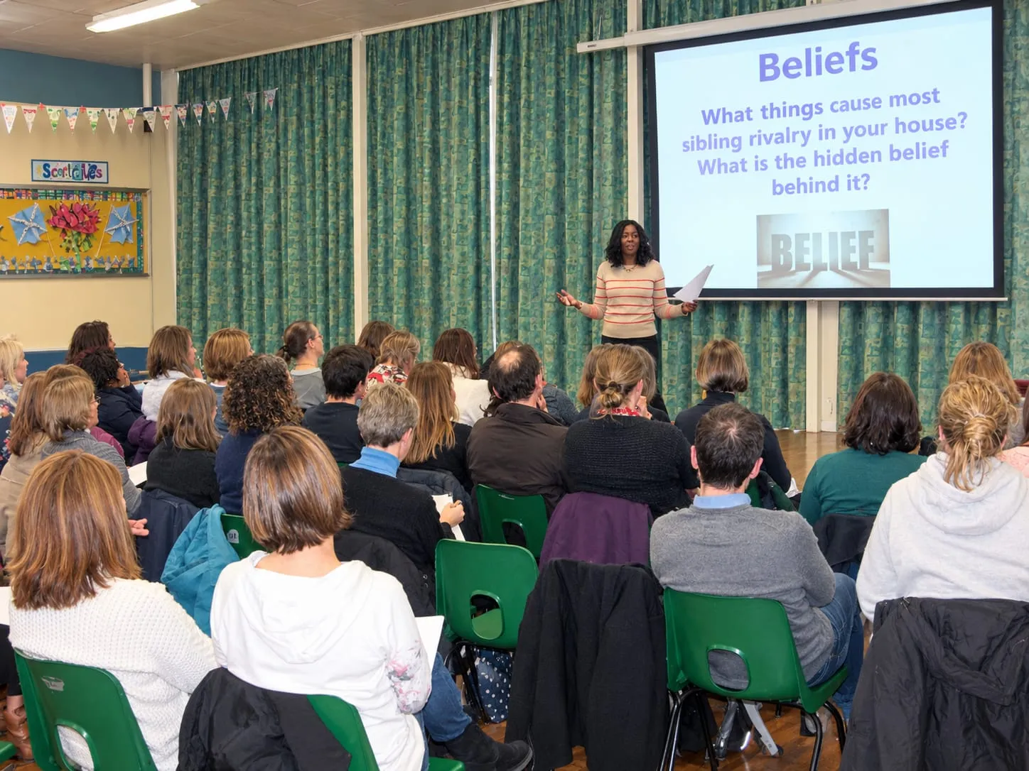 Lorraine Lee speaking to a hall of parents at a primary school, beside a slide about beliefs
